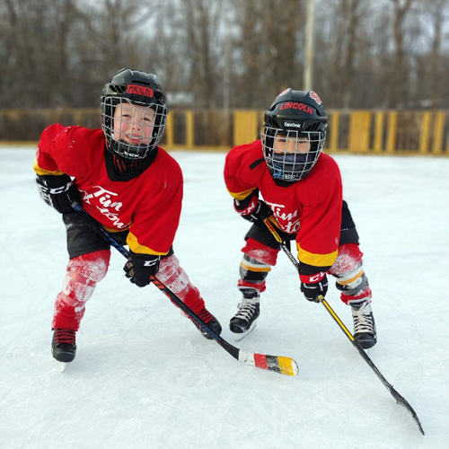 Two smiling children in hockey gear with red and yellow jerseys on a skating rink.